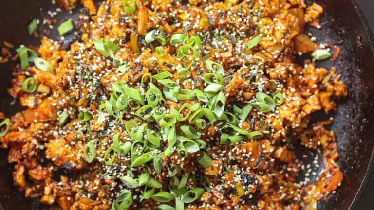 A close-up of vegan kimchi fried rice in a skillet, topped with sliced green onions, sesame seeds, and crumbled nori. The dish has a vibrant orange hue from the kimchi and is richly garnished, making it visually appealing.